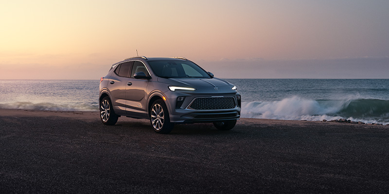 A silver Buick Encore GX parked on a beach at sunset, with the ocean waves in the background, showcasing its sleek and modern design.