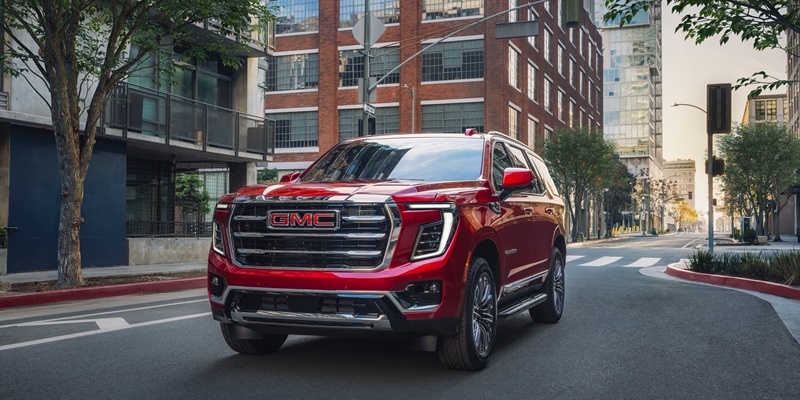 A red GMC Yukon driving through a city street, showcasing its bold design with a prominent front grille and modern features, surrounded by urban buildings.
