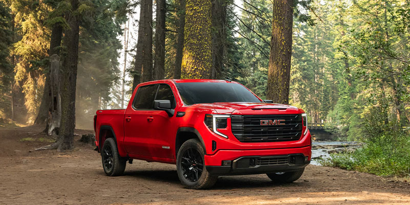Red GMC pickup truck parked on a dirt road in a forested area with tall trees and a creek visible in the background.