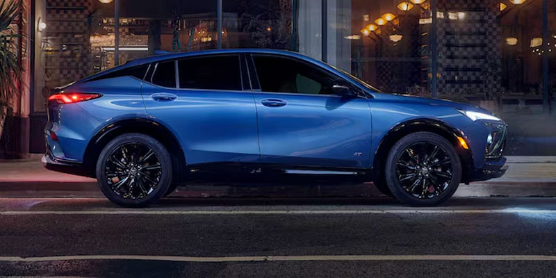 Side profile view of a blue modern crossover SUV parked on a city street at night, with illuminated storefront lights reflecting on its body and wheels.
